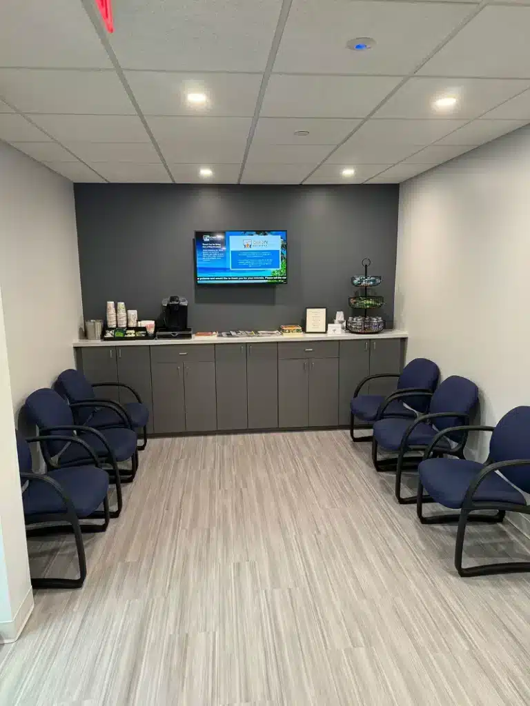Patient waiting room with blue chairs, a wall-mounted TV, and a refreshment counter at ProHealth Chiropractic Rockville clinic