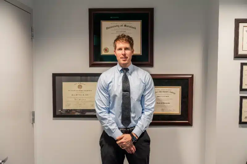 Portrait of a chiropractor standing in front of his framed university and chiropractic college diplomas at ProHealth Chiropractic Rockville clinic