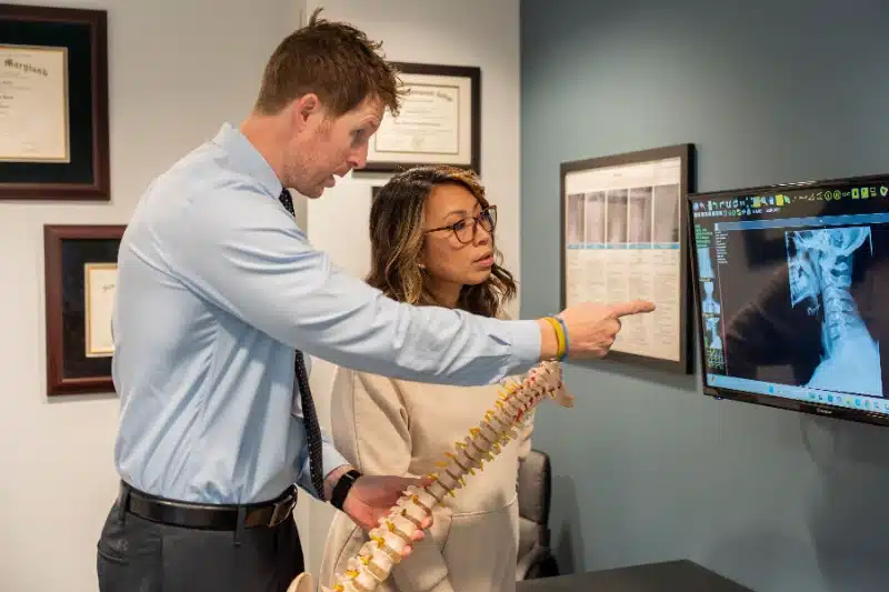 Chiropractor holding a spine model while showing a patient a neck X-ray on a monitor at ProHealth Chiropractic Rockville clinic